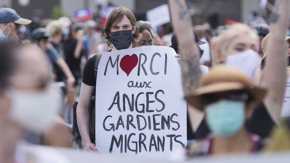 Une femme tenant une pancarte sur laquelle il est inscrit : « Merci aux anges gardiens migrants ».
