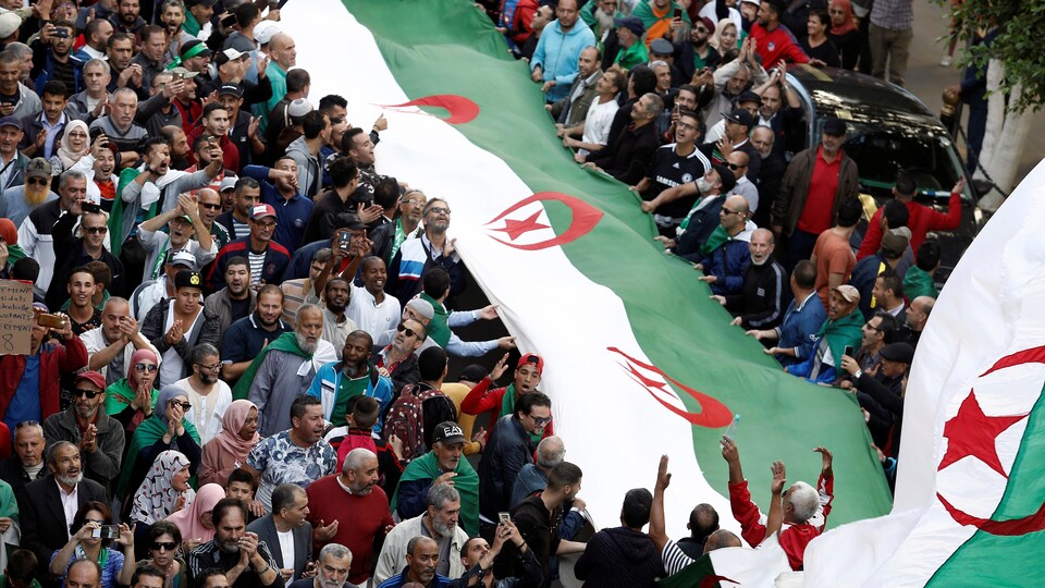Des centaines de manifestants tiennent un long drapeau algérien dans la rue.