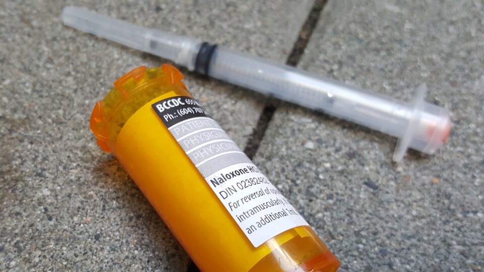 Une seringue usée et un flacon ouvert sur lequel est apposé une ordonnance pour Naloxone sont sur le pavé.