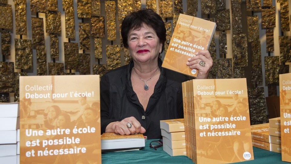 Suzanne-G. Chartrand tenant une copie du livre « Une autre école est possible et nécessaire » devant une table où se trouvent plusieurs autres copies du livre.