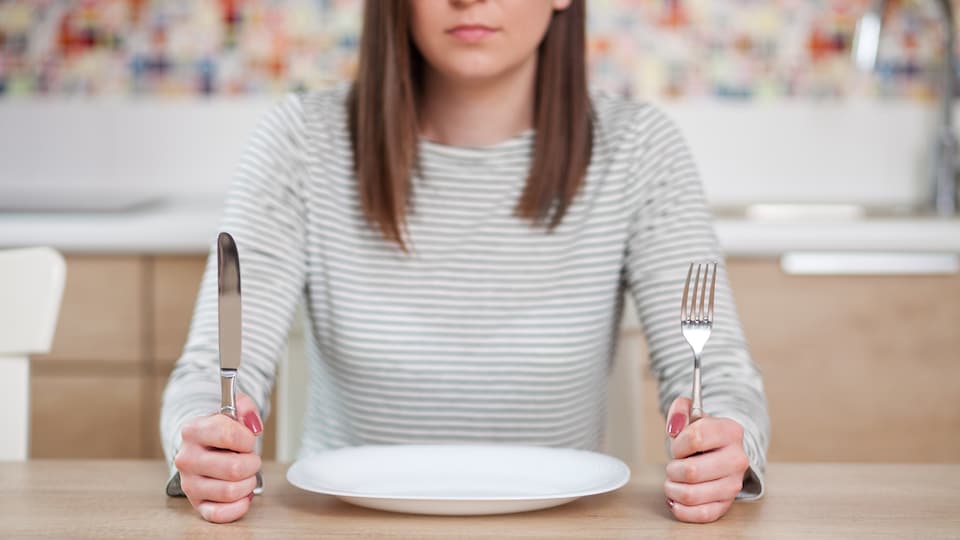 Une jeune femme mécontente est assise devant une assiette vide et tient des ustensiles dans ses mains.
