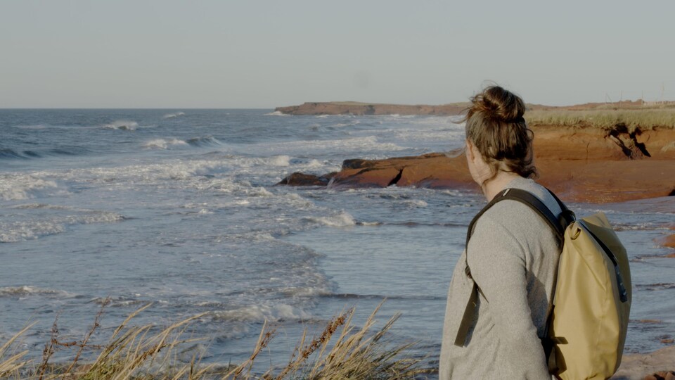 Une femme regarde l'horizon. Elle est au bord de la mer. On peut voir des falaises rouges en arrière-plan.