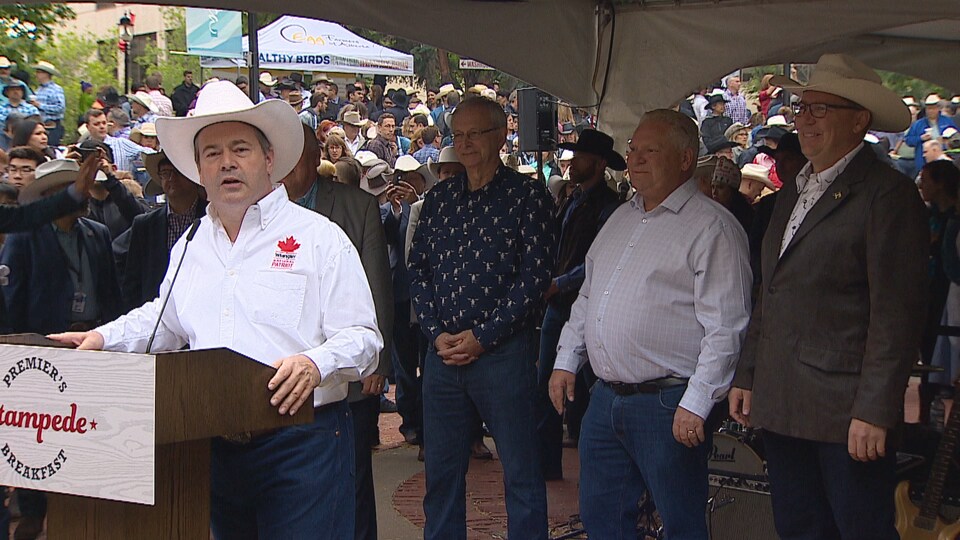 Jason Kenney parle a un micro debout devant une foule tandis que les premiers ministres Doug Ford, Blaine Higgs, Scott Moe sont debout derrière lui.
