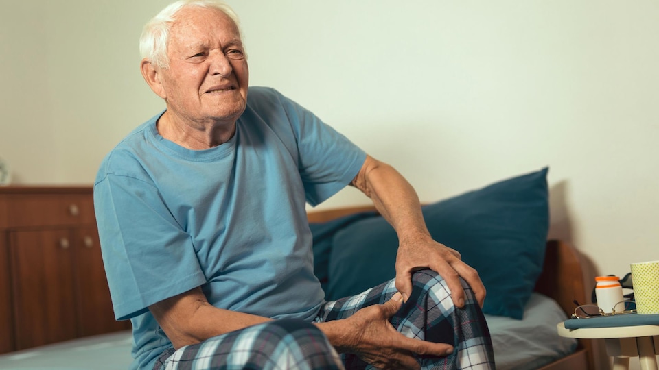 Un homme âgé grimace de douleur en se tenant le genou gauche. Il est assis sur un lit simple.