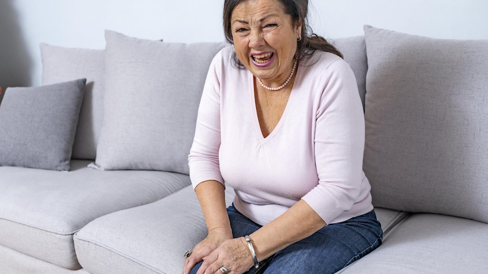 Une femme âgée grimace de douleur en se tenant le genou droit. Elle est assise sur un sofa.