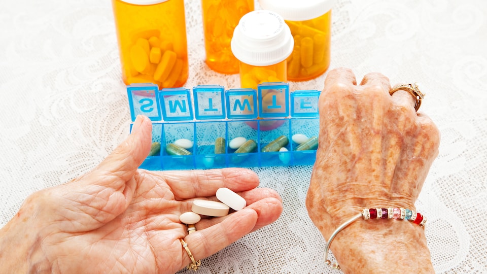 Les mains d'une vieille dames tiennent des médicaments variés. En arrière-plan, on voit un pilulier et des contenants de médicaments. 