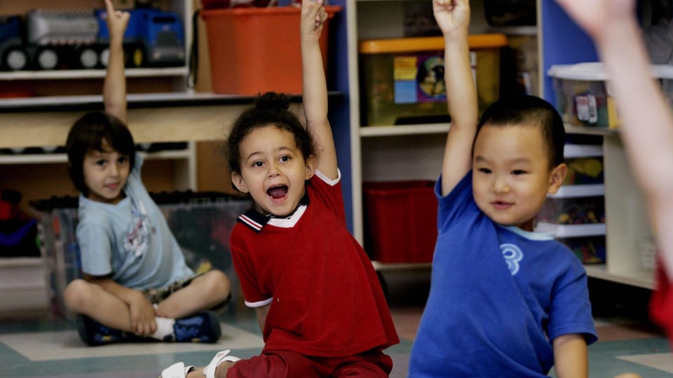 Trois jeunes enfants de diverses nationalités sont assis au sol et lèvent leur main dans une salle de classe.