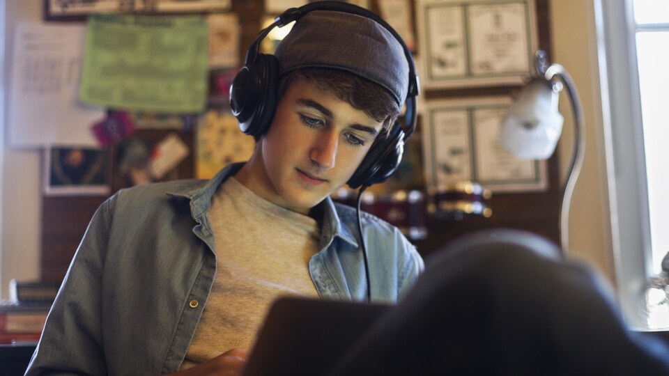 Un adolescent travaille sur son ordinateur avec des écouteurs sur ses oreilles, dans sa chambre. 
