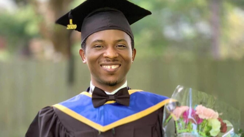 Un homme souriant habillé d'une toge et d'un mortier tenant dans ses mains un diplôme et des fleurs.