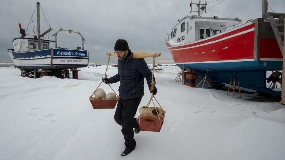 Un homme porte un joug et du bois à travers des bateaux sortis de l'eau pour l'hiver aux Îles-de-la-Madeleine.