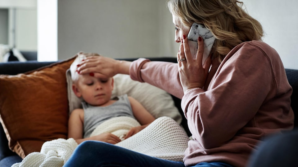 Une mère met une main sur le front de son fils pour prendre sa température, tout en portant son téléphone à l'oreille de l'autre main. 