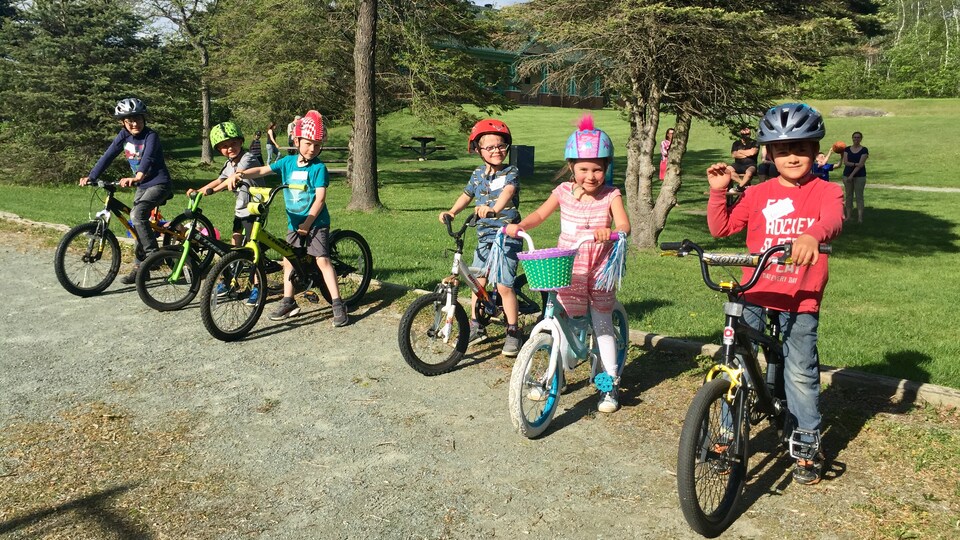 Six enfants, cinq garçons et une fille, à cheval sur leur bicyclette. Ils portent tous un casque. Ils sont dans un parc sur un sol rocheux. Ils sont arrêtés, les pieds au sol, et regardent la caméra.