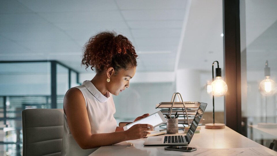 Une femme travaille sur une tablette informatique; un ordinateur est posé sur son bureau.
