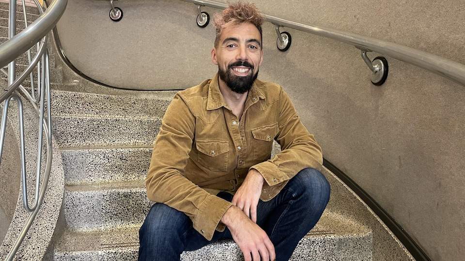 Eric Plamondon, souriant, est assis sur les marches d'un escalier des locaux de CBC/Radio-Canada au Manitoba