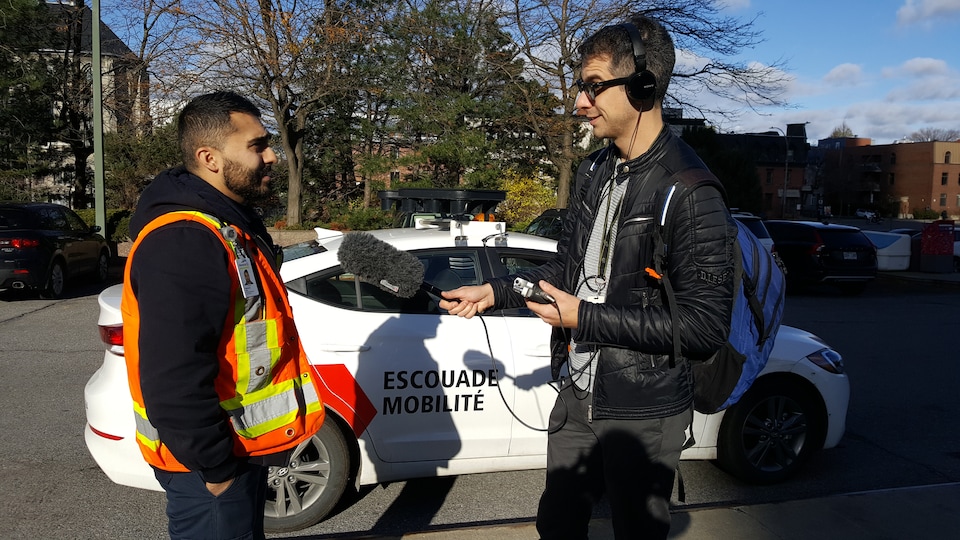 Un homme portant un dossard orange parle dans un micro tenu par un autre homme, devant une voiture sur laquelle on peut lire « Escouade mobilité ».