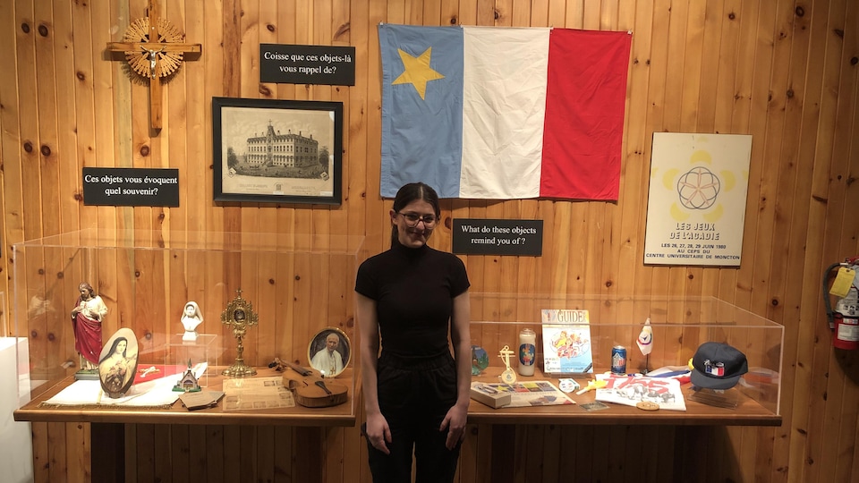 Une femme est debout devant une exposition de différents objets acadiens et religieux.