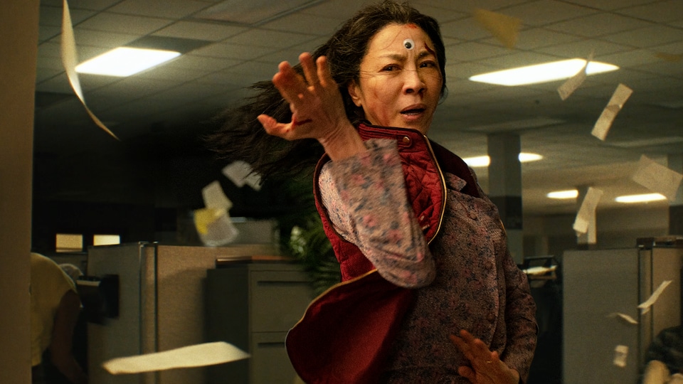 L'actrice place ses bras en position de combat dans un bureau et des reçus de caisse volent autour d'elle. 