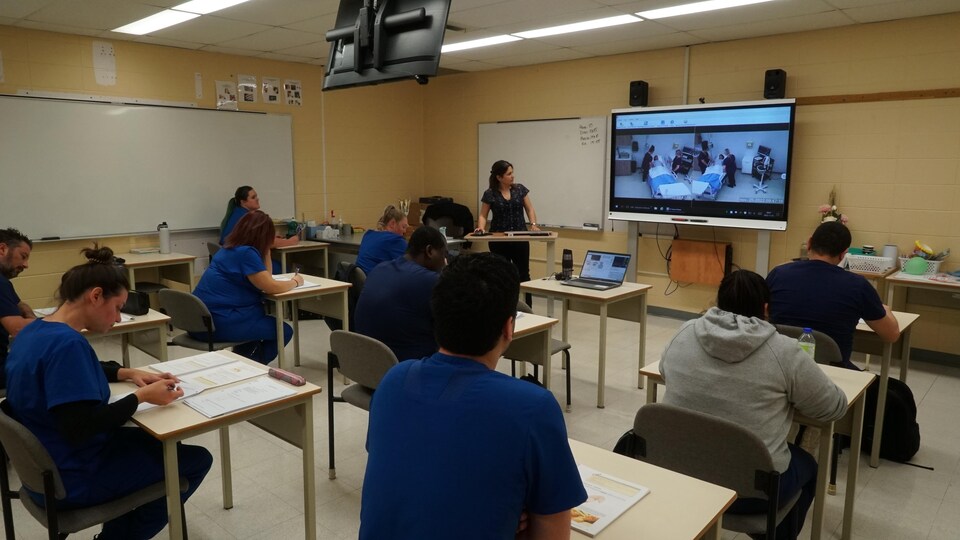 Des étudiants regardent un écran vidéo en avant de la classe. Sur l'écran, on peut apercevoir deux infirmières qui sont près d'un mannequin intelligent qui joue le rôle d'un patient dans un hôpital.