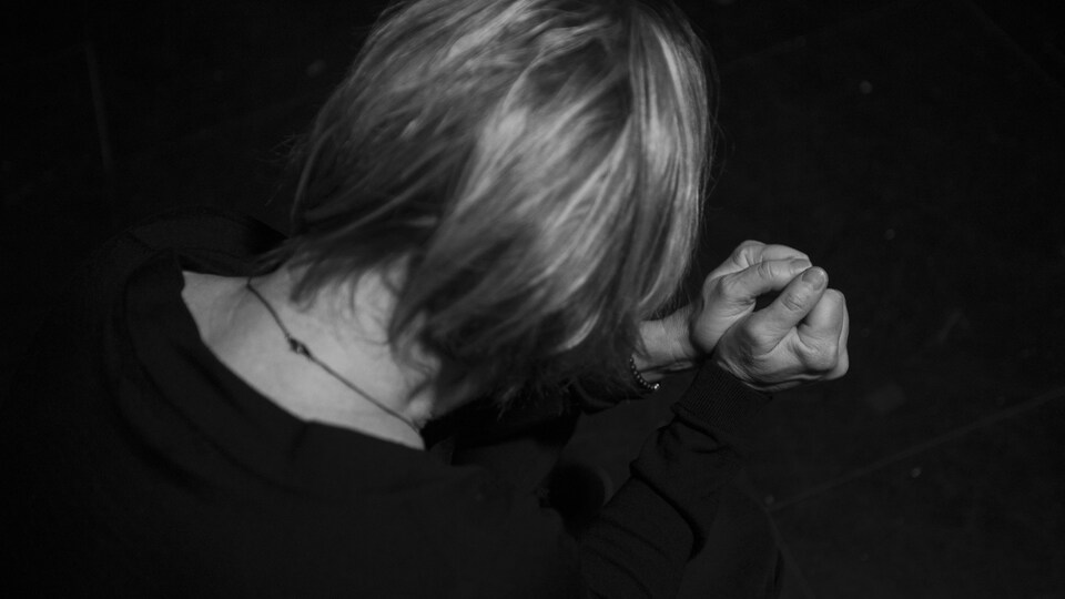 Dans un fond sombre, une femme vue de dos semble faire une prière, sauf qu'elle a les poings serrés.