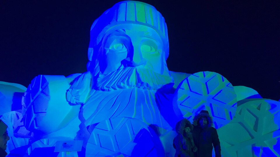 Deux personnes se font prendre en photo devant une sculpture de glace d'un homme barbu avec une tuque.