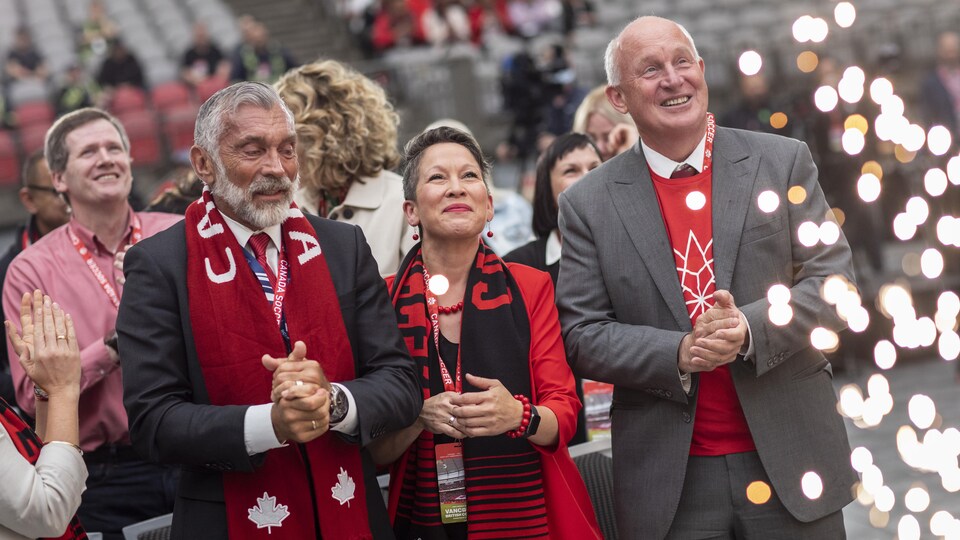 3 personnes en portrait devant une foule, dans un cadre manifestement festif, avec des écharpes du Canada.