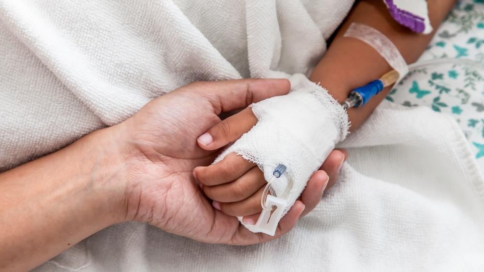 Un homme tient la main d'un enfant couché sur un lit d'hôpital et branché à un soluté.