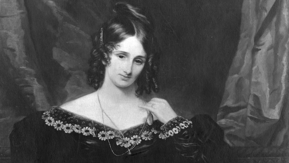 Mary Shelley, une plume à la main, semble écrire sur une feuille alors que plusieurs livres sont éparpillés sur la table.