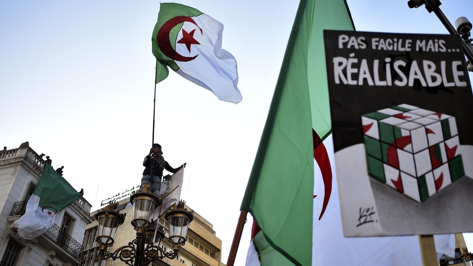 Un jeune homme juché sur un lampadaire tenant un drapeau de l'Algérie. Ainsi qu'une pancarte sur laquelle il est inscrit : « Pas facile mais... réalisable » avec un gros cube Rubik aux couleurs du drapeau algérien.