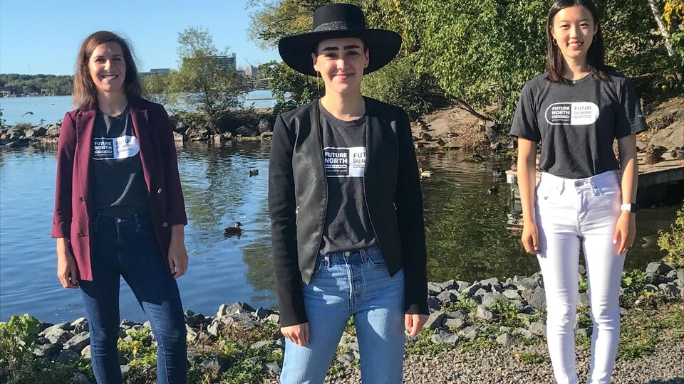 Trois jeunes femmes sourient devant un lac à Sudbury en portant les chandails de Futur du Nord.