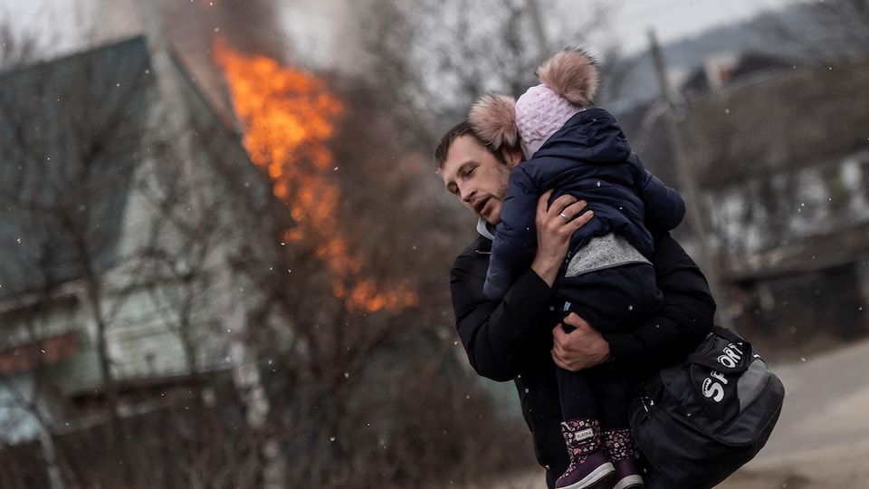 un homme tient un enfant, une maison en feu en arrière plan.