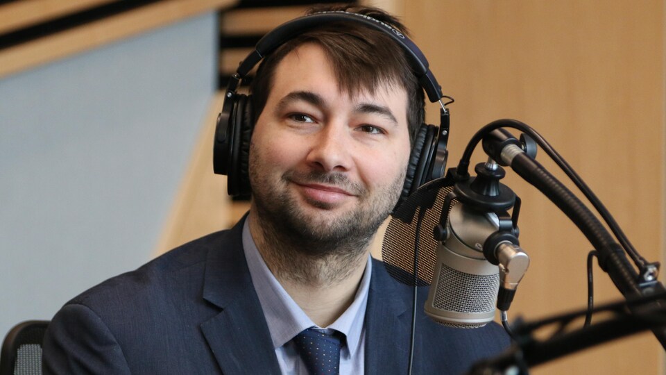 Un homme qui porte un casque d'écoute sur la tête sourit derrière un micro dans un studio de radio.