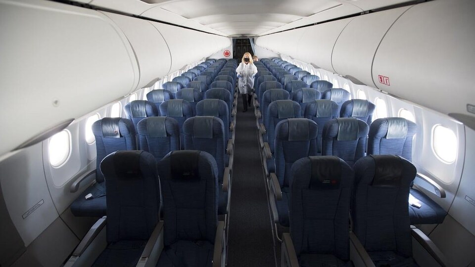 Une femme marche dans l'allée centrale d'un avion d'Air Canada. Elle porte un masque, un sarrau blanc et des gants.
