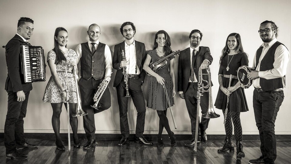 Photo noir et blanc de huit musiciens, trois femmes, cinq hommes: accordéoniste, tromboniste, trompettiste, clarinettiste, violoniste, flûtiste, percussionniste.