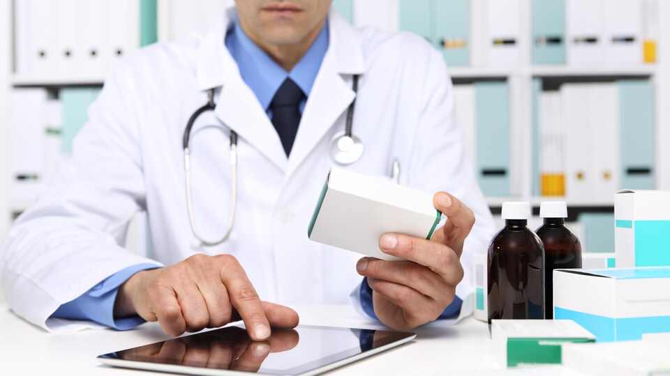 Un médecin tenant une boîte de médicaments et consultant une tablette électronique.