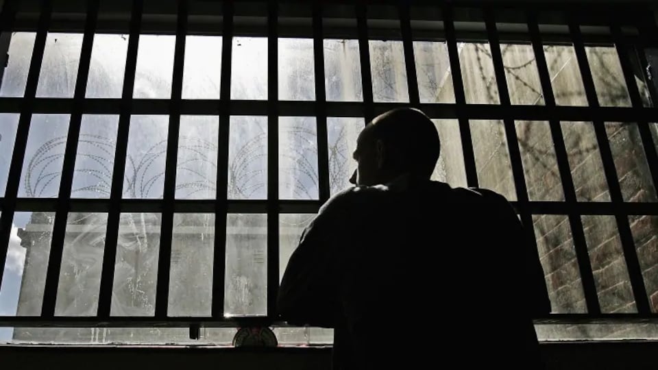 Un détenu regarde par une fenêtre doublée de barreaux dans une prison.