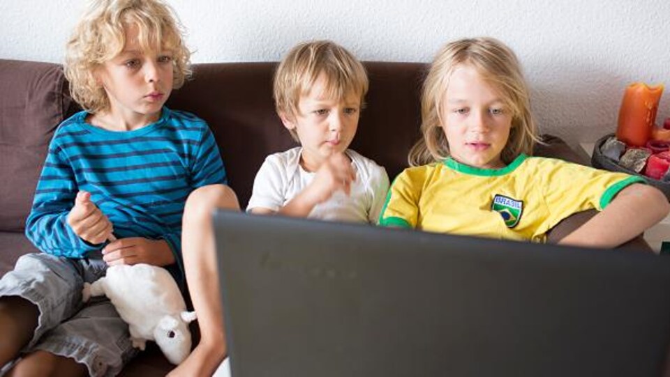 Three young children sitting on a couch look at the screen of a laptop computer. 