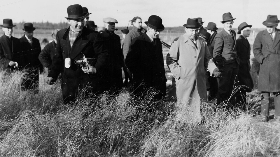 Sur une ferme à l'extérieur, un groupe d'hommes vêtus en veston-cravate et chapeau haute-forme. Parmi eux, l'abbé Maurice Proulx à l'avant-plan avec une caméra sous le bras.
