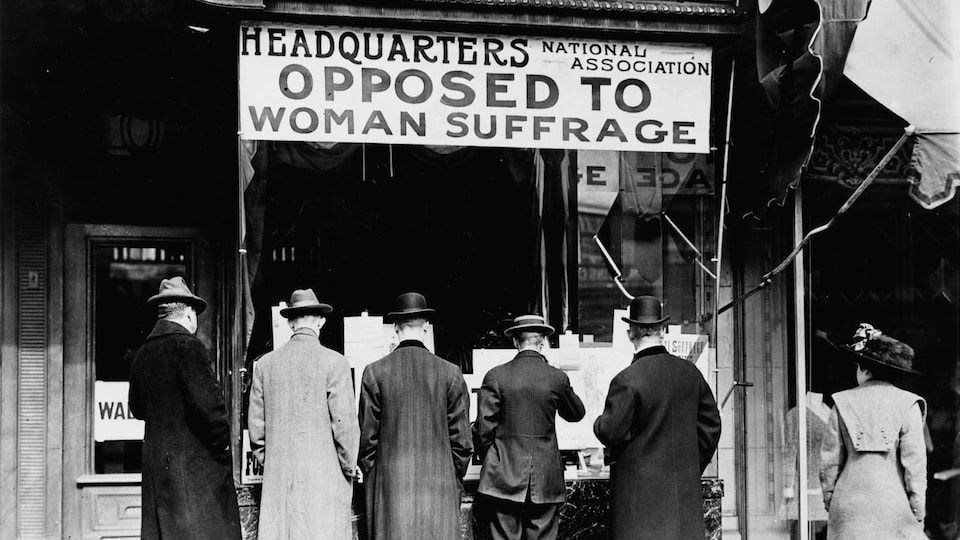 Cinq hommes de dos regardent la devanture du siège de l’association opposée au suffrage des femmes, alors qu'une passante les regarde.