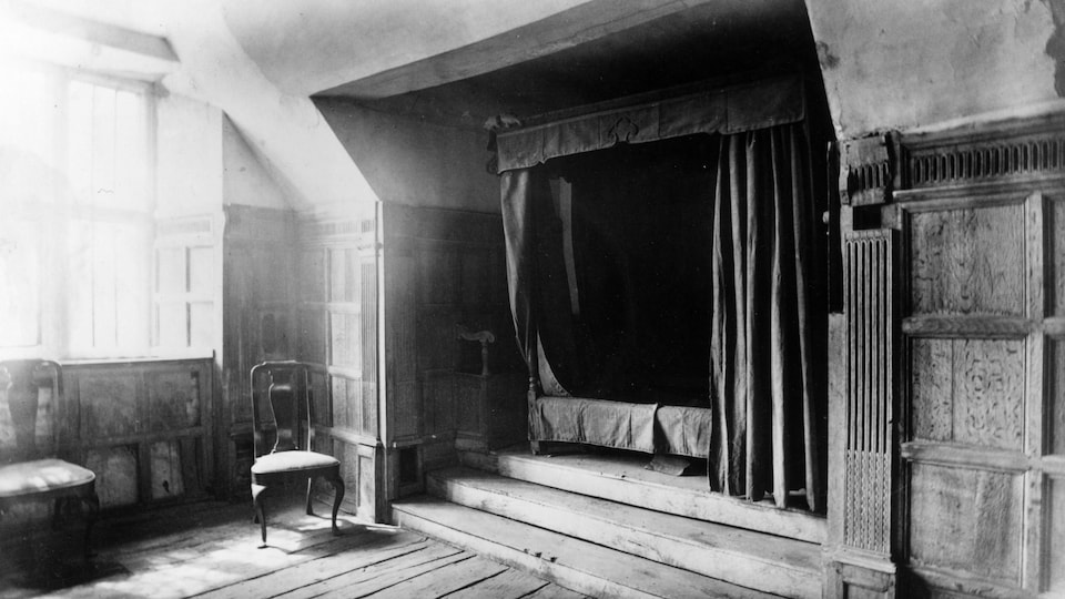 Dans une chambre, un lit est située dans une alcôve drapée.