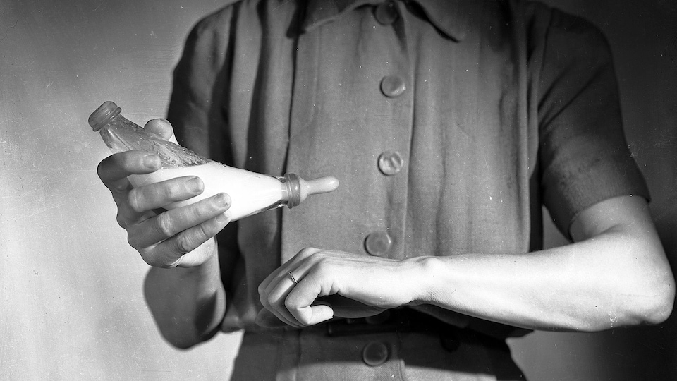 Photo en noir et blanc d'une femme qui vérifie la température du lait d'un biberon en versant une goutte sur sa main.