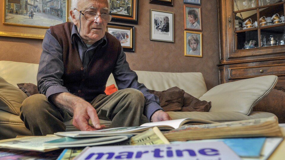 L’illustrateur Marcel Marlier est assis sur un divan et feuillette un livre de  la série Martine.