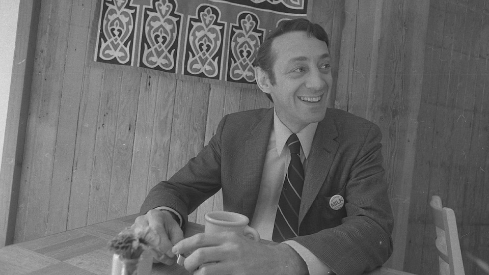 Photo en noir et blanc montrant le militant Harvey Milk souriant, assis à la table d'un restaurant.