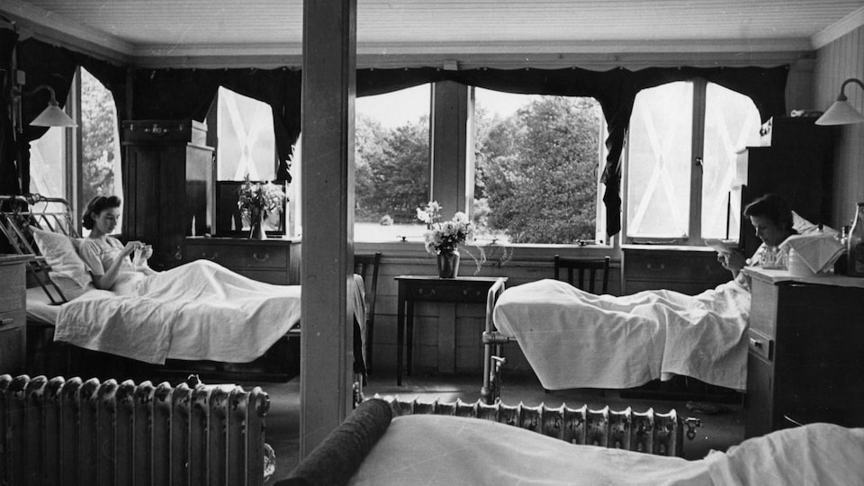 Photo en noir et blanc montrant deux femmes dans des lits drapés de blanc à l'intérieur d'une pièce élégamment décorée.