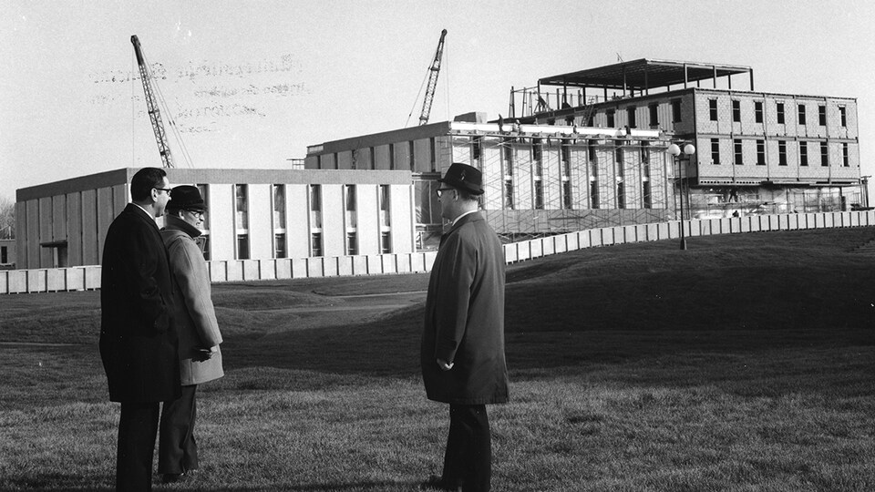 Trois hommes regardent l'Université de Moncton en construction.