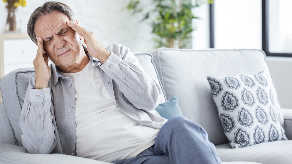 Assis sur un divan, un homme se touche les tempes en raison d'un mal de tête.
