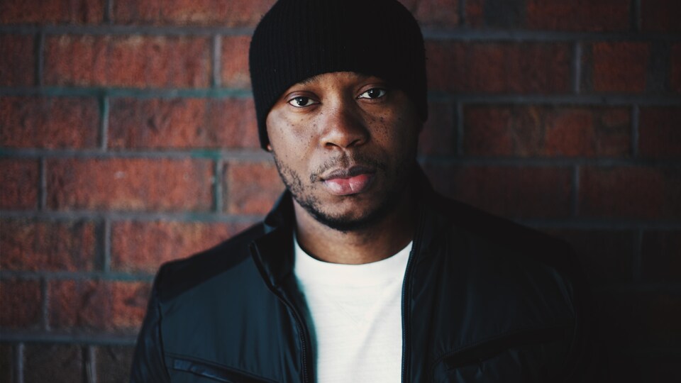 L'artiste portant un chandail blanc ainsi qu'une tuque et un manteau noirs.