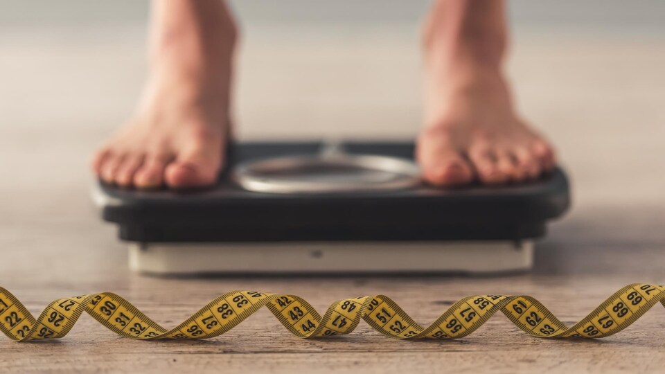 Une photo montrant les pieds d'une personne sur une balance. En avant-plan, un ruban à mesurer est visible.