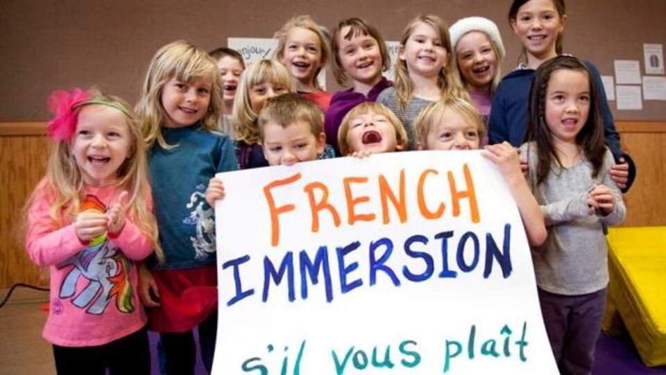 Une douzaine d'enfants de 6-7 ans sourient et et tiennent une pancarte sur laquelle il est écrit: French immersion s'il-vous-plaît.