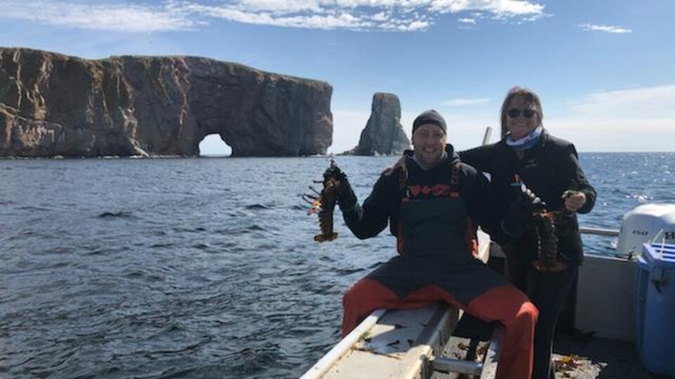 Jimmy Lepage assis sur navire de pêche devant le rocher Percé tient un homard dans chaque main en compagnie d'une femme.