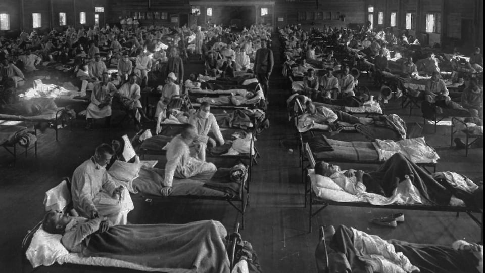 Des dizaines de patients sont alités sur des lits placés en rangée dans une salle aux dimensions importantes qui s’apparente à un auditorium. Des médecins et des infirmières se tiennent au chevet des malades. La photographie est en noir et blanc.